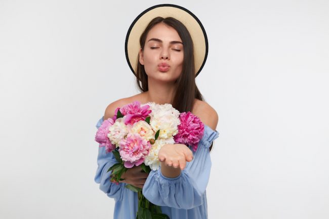 Portrait of attractive, nice looking girl with long brunette hair. Wearing a hat and blue dress. Holding a bouquet of flowers and sending a kiss. Stand isolated over white background